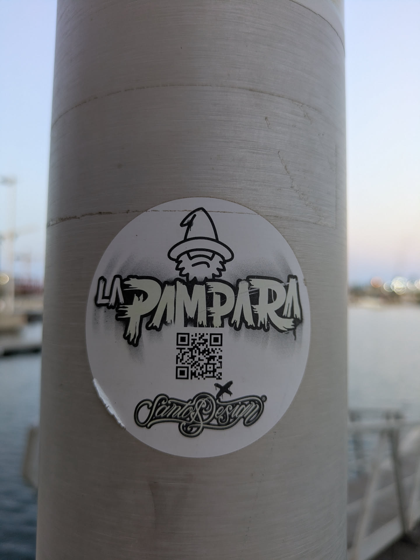 A round white sticker of the band La Pampara is stuck on a grey pole. The sticker features a logo of a wizard above the band name, a QR code, and the name Santos Design.