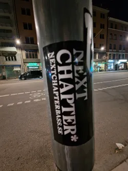 A black and white sticker on a metal pole with the text "NEXT CHAPTER*" and the website "NEXTCHAPTERBASS.SE" underneath. The sticker is rectangular and has a slightly worn look.