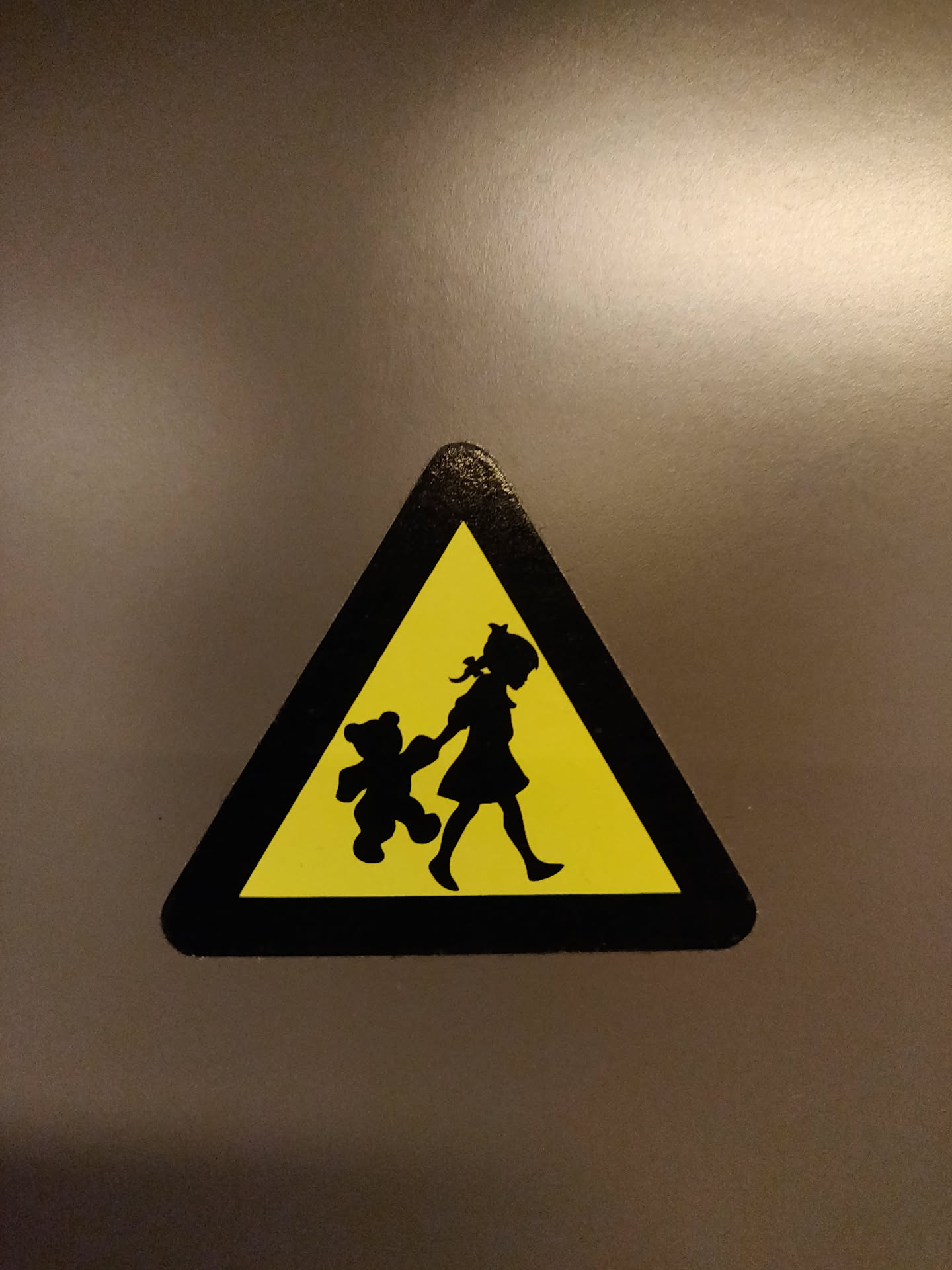 This sticker is in the shape of a triangle and depicts a silhouette of a girl walking with her teddy bear.  It has a yellow background and black border, mimicking a warning sign. 