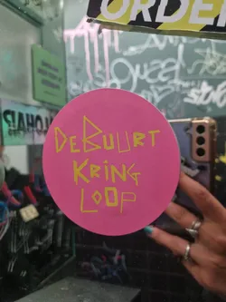 Pink round sticker with yellow text on it. The text says "Debuurt Kring Loop".