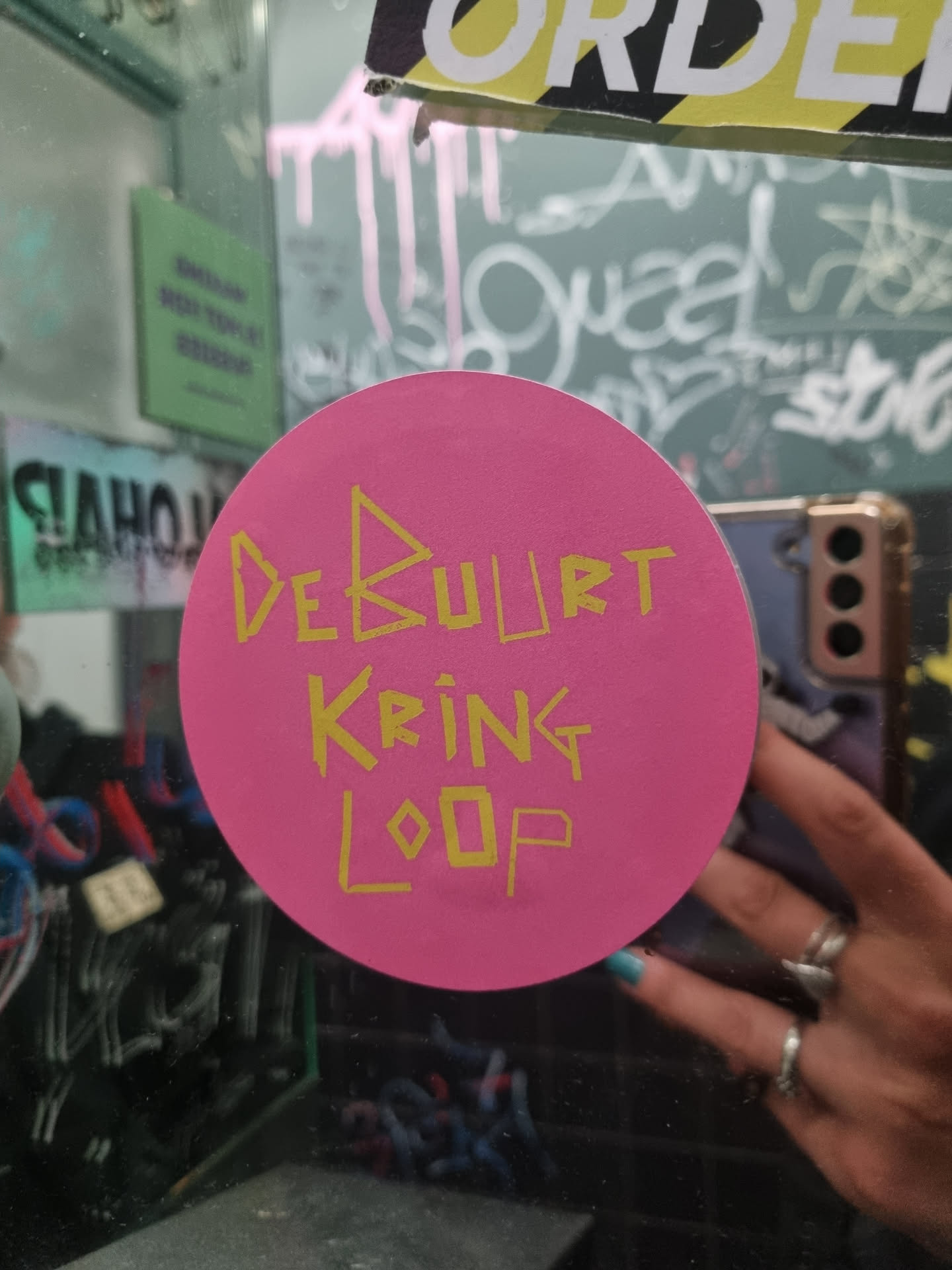 Pink round sticker with yellow text on it. The text says "Debuurt Kring Loop".