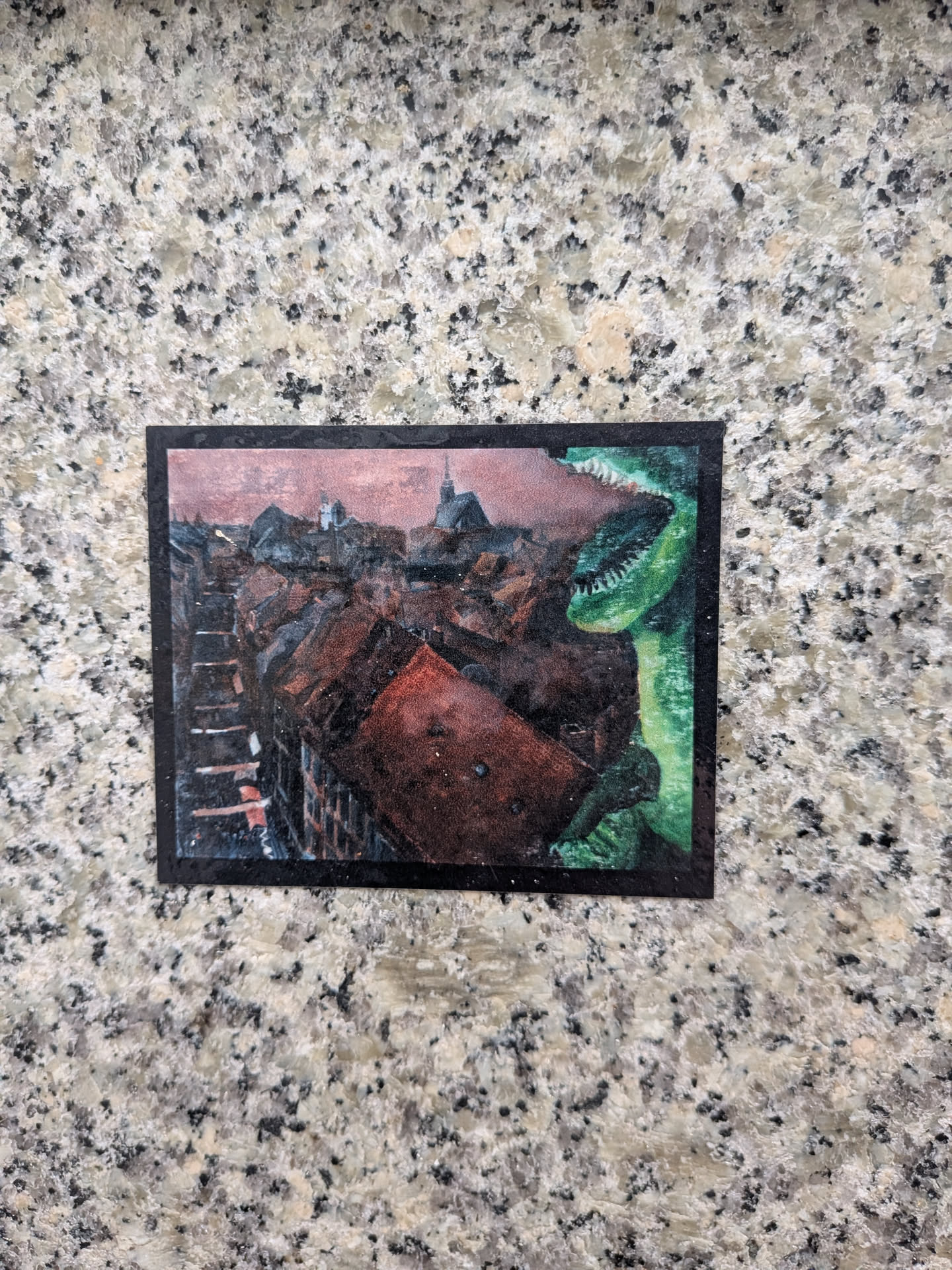A rectangular sticker bordered in black featuring a painted scene. An aerial view reveals a European old town with red-tiled rooftops stretching towards a church spire beneath a dusky pinkish-grey sky. On the right side, a colossal green dinosaur with sharp teeth and open jaws peers menacingly over the cityscape.