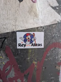 A rectangular white sticker featuring a cartoon rooster dressed in royal attire, including a small crown, cape, and holding a scepter. The character is set inside a blue circle above the text 'El Rey de las Alitas', with 'El' and 'de las' written in orange and 'Rey' and 'Alitas' in bold black letters.