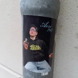 A sticker featuring a photo of a young man named Aasi, wearing a black hoodie with yellow text that says "VOLTUE HUSLIES", and holding a can of drink. The background is black and the text "Aasi 245" is written in a stylized font in the upper right corner.
