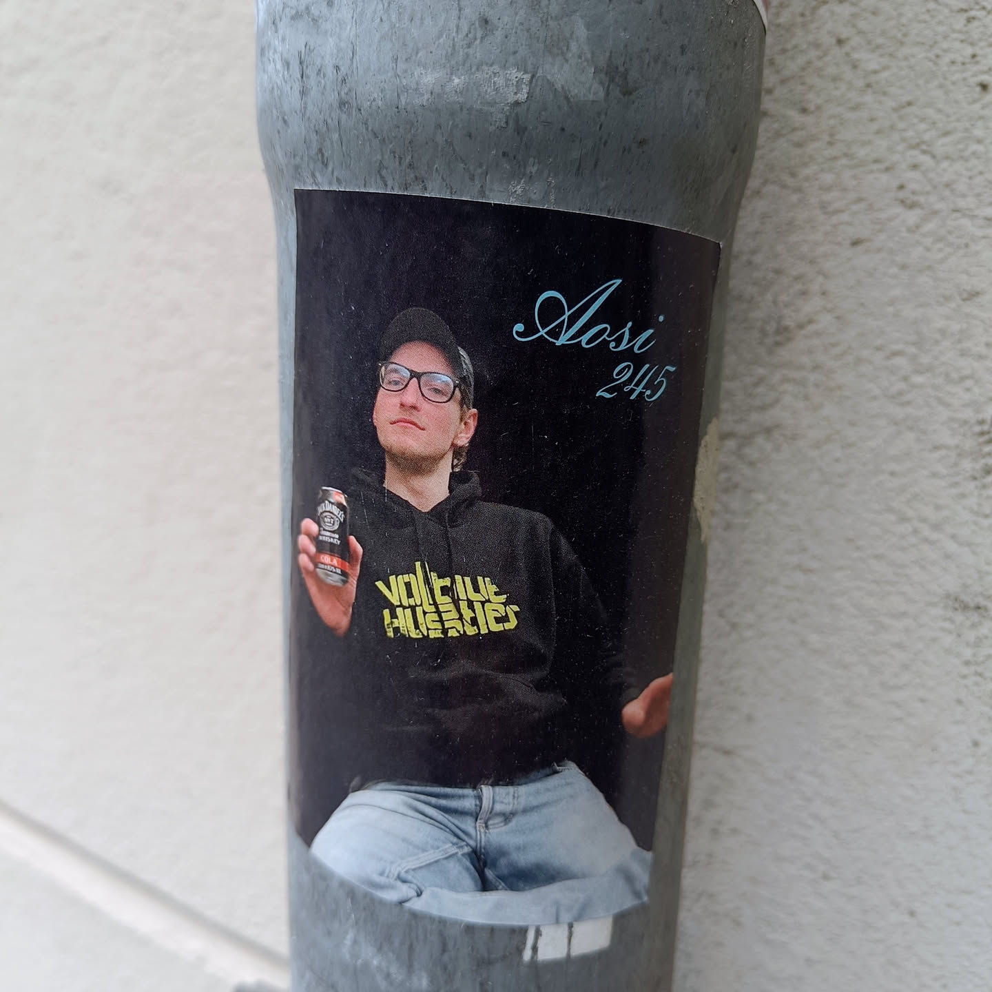 A sticker featuring a photo of a young man named Aasi, wearing a black hoodie with yellow text that says "VOLTUE HUSLIES", and holding a can of drink. The background is black and the text "Aasi 245" is written in a stylized font in the upper right corner.