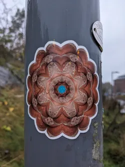 This sticker shows a mandala with copper tones and a teal center. It's placed on a gray metal pole, and water droplets are visible on its surface. The mandala has a detailed, almost fractal-like pattern.