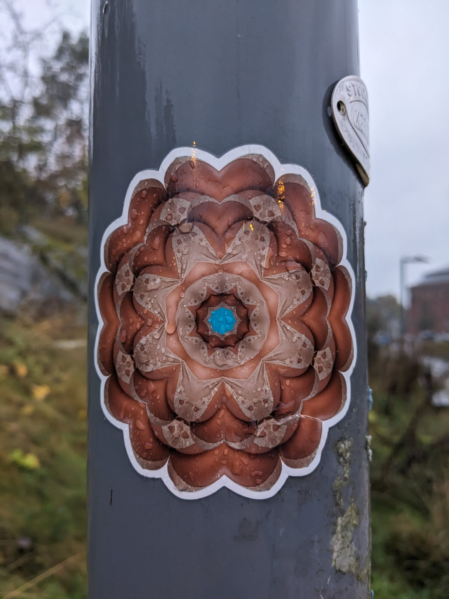 This sticker shows a mandala with copper tones and a teal center. It's placed on a gray metal pole, and water droplets are visible on its surface. The mandala has a detailed, almost fractal-like pattern.