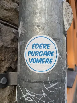 A round sticker with the words "EDERE PURGARE VOMERE" written in a stylized blue font on a white background. The sticker is affixed to a weathered metal pole.