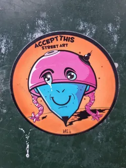 Round sticker featuring an illustration of a crying cartoon character that resembles a spinning top with a pink helmet and pink braids. The character has a blue body and a happy face. The background is orange. The text "ACCEPT THIS STREET ART" is written at the top of the sticker. The artist's signature "KRS-b" is visible at the bottom. 
