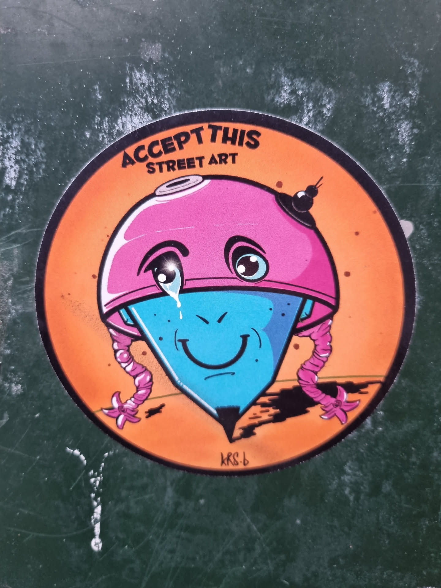 Round sticker featuring an illustration of a crying cartoon character that resembles a spinning top with a pink helmet and pink braids. The character has a blue body and a happy face. The background is orange. The text "ACCEPT THIS STREET ART" is written at the top of the sticker. The artist's signature "KRS-b" is visible at the bottom. 