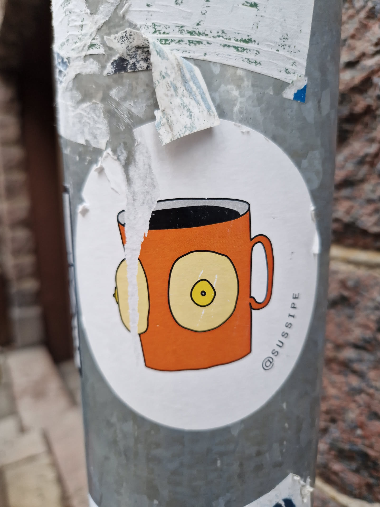 A sticker of an orange coffee mug with two breasts on the side. The artist's Instagram handle is @sussipe.