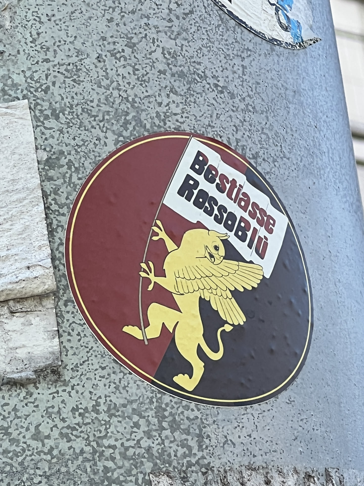 A circular sticker featuring a golden winged lion standing on its hind legs, holding a white flag. The background is split vertically, with the left half in dark red/maroon and the right half in dark blue/black. A thin golden-yellow border encircles the design. The lion has a fierce expression and prominent wings. The flag it holds displays the text 'Bestiasse' in black and 'RossoBlü' in red.
