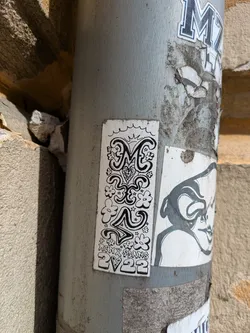A black and white sticker adhered to a gray pole. The sticker features decorative text and floral elements in a doodle style. The text reads "Monx Spans 2V22".