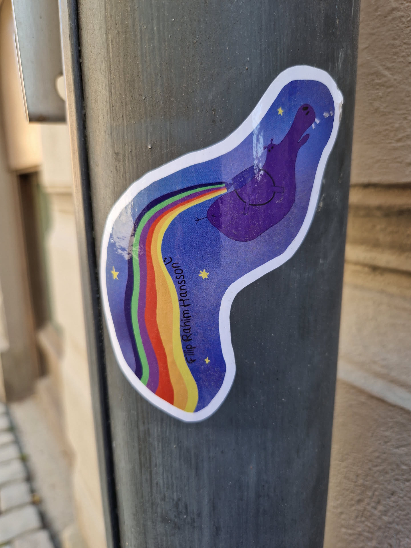 A sticker featuring a purple hippopotamus on a blue background pooping a rainbow. The text 'Filip Rahim Hansson' is written on the sticker.