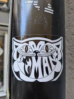A black and white sticker featuring a stylized cat face with the word "Mad" written inside. The cat has large, oval eyes and a mischievous grin. The style is reminiscent of vintage cartoons or illustrations.