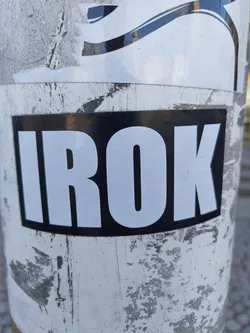 A black and white sticker with the word "IROK" in bold, uppercase, white lettering on a black background. The sticker is affixed to a worn, light-grey surface with visible scratches and other stickers underneath. There are no visible social media links or QR codes on the sticker itself.