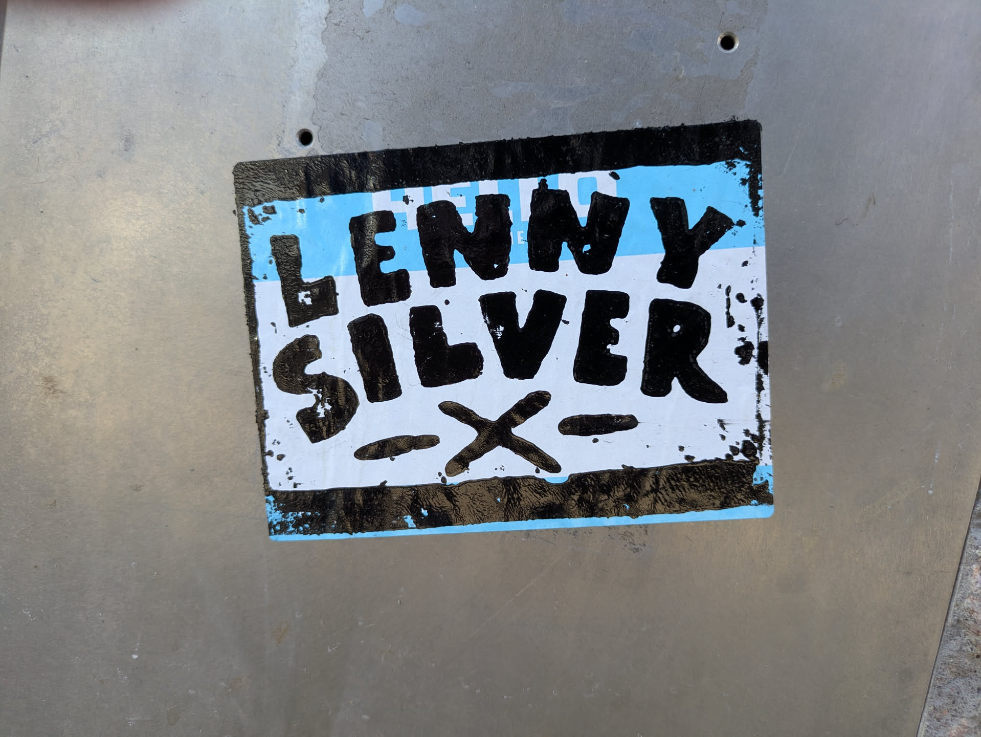 A rectangular sticker with a black border, featuring a light blue background. Prominently displayed in the center are the words "LENNY" and "SILVER" in thick, black, hand-drawn block letters. Below "SILVER", there is a stylized black 'X' with two horizontal dashes on either side of it. The edges of the sticker show signs of wear and tear, with some black paint splatter and peeling.