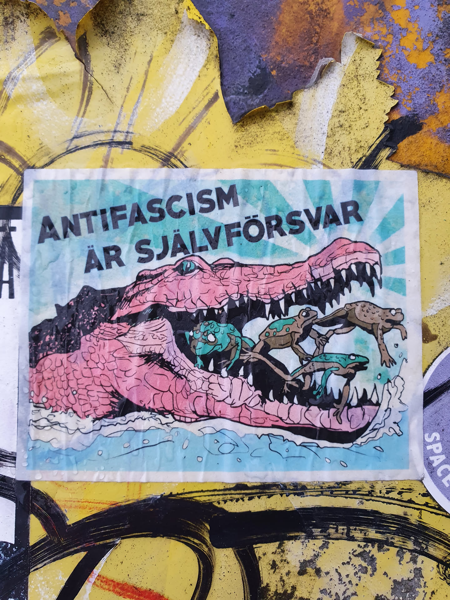 A sticker with an illustration of a crocodile with frogs in its mouth. The text on the sticker says "Anti-fascism är självförsvar" which translates to "Anti-fascism is self-defense" in Swedish.