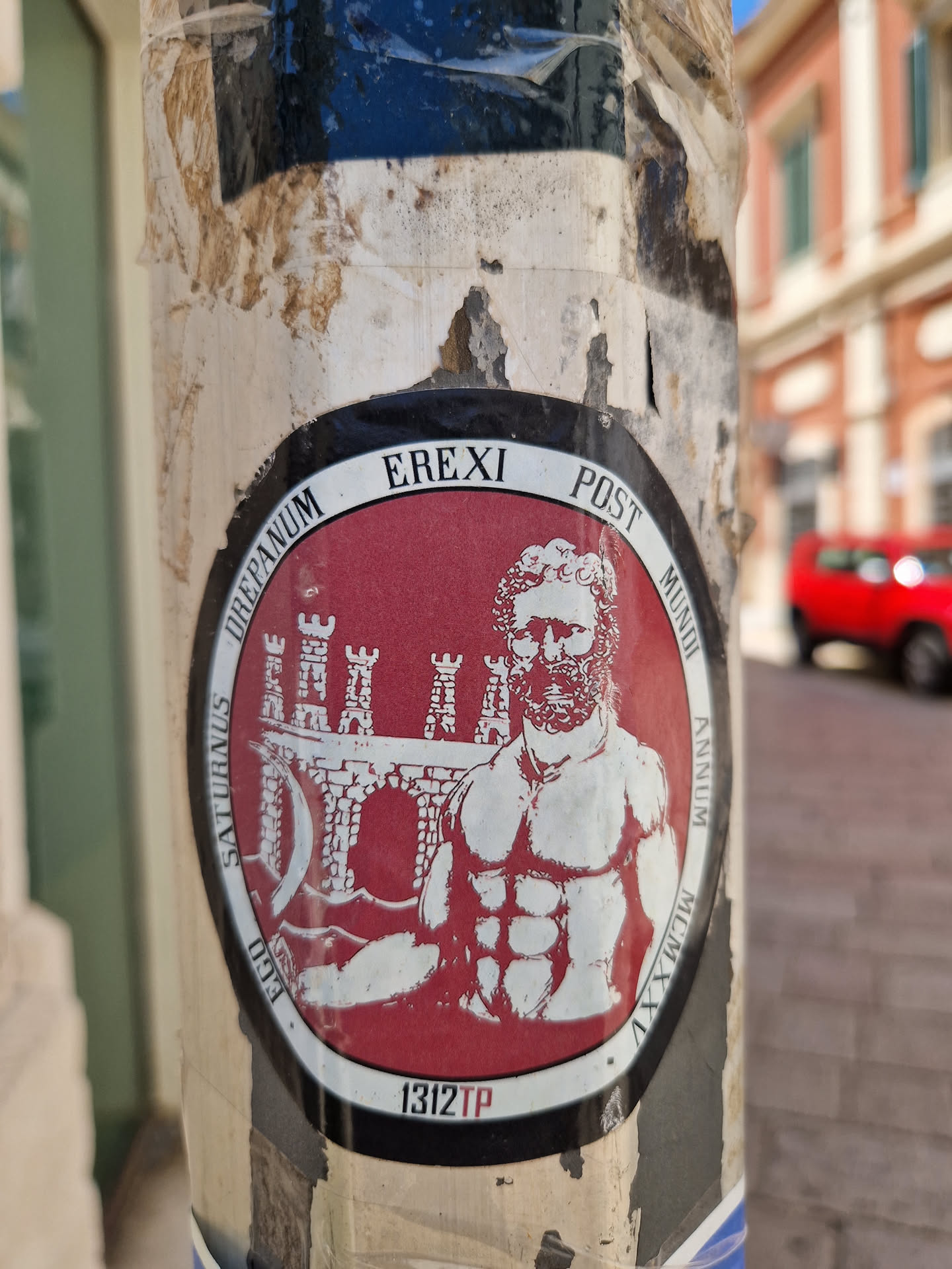 This round sticker features a red background with a white illustration of a muscular man with a beard who appears to be inspired by Roman iconography. He is situated in front of a structure that resembles a fortress or city gate. The surrounding text suggests a reference to a historical event or era. The number "1312TP" is printed at the bottom.