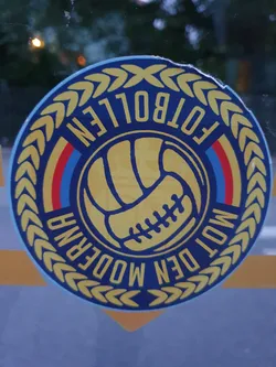 Round sticker featuring a stylized football and the text "Modern Fotbollen" within a laurel wreath. The colors are predominantly blue, yellow, and red.