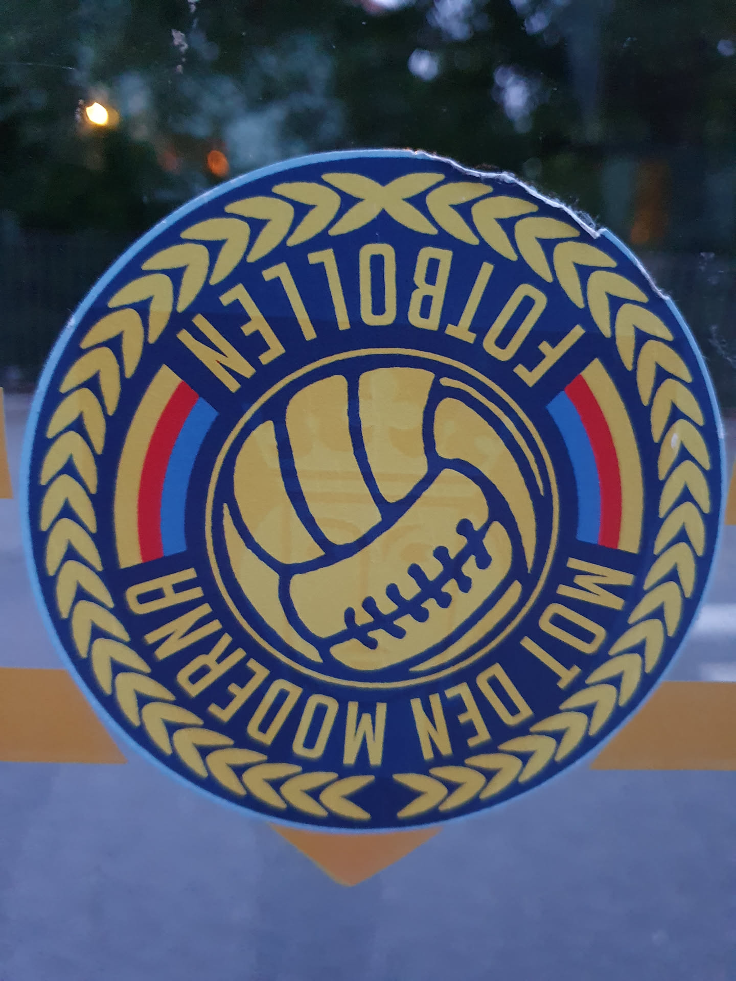 Round sticker featuring a stylized football and the text "Modern Fotbollen" within a laurel wreath. The colors are predominantly blue, yellow, and red.