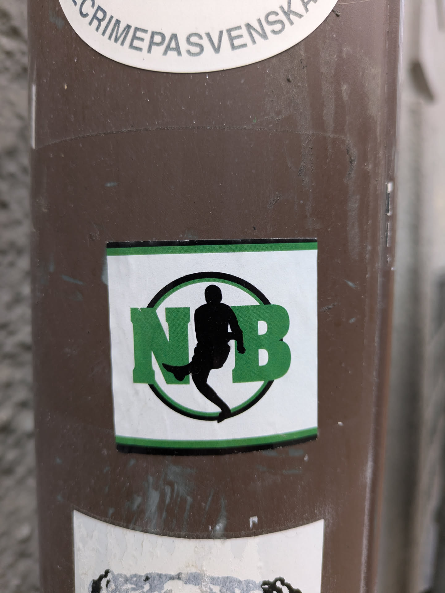 A close-up shot of a sticker featuring the letters "NB" in green, encircled within a black circle.  A black silhouette of a person in a dynamic sports pose is situated between the letters. The sticker has a white background and a dark green border. It is affixed to a brown pole.
