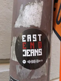 The sticker has the text "EAST END JEANS" stacked vertically, with "END" in red. Below the text is a Spotify code. It is placed on a weathered brown pole.