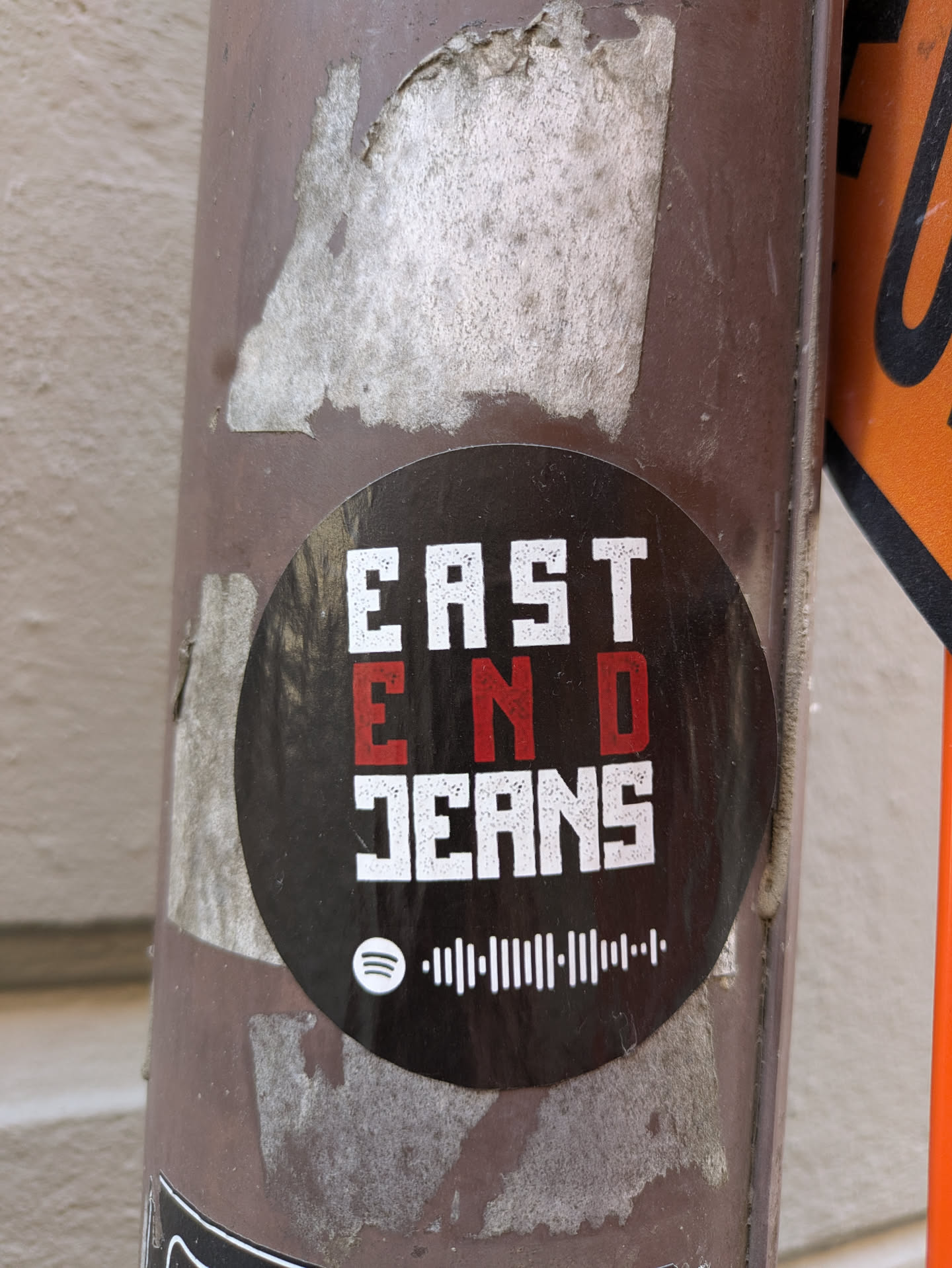 The sticker has the text "EAST END JEANS" stacked vertically, with "END" in red. Below the text is a Spotify code. It is placed on a weathered brown pole.