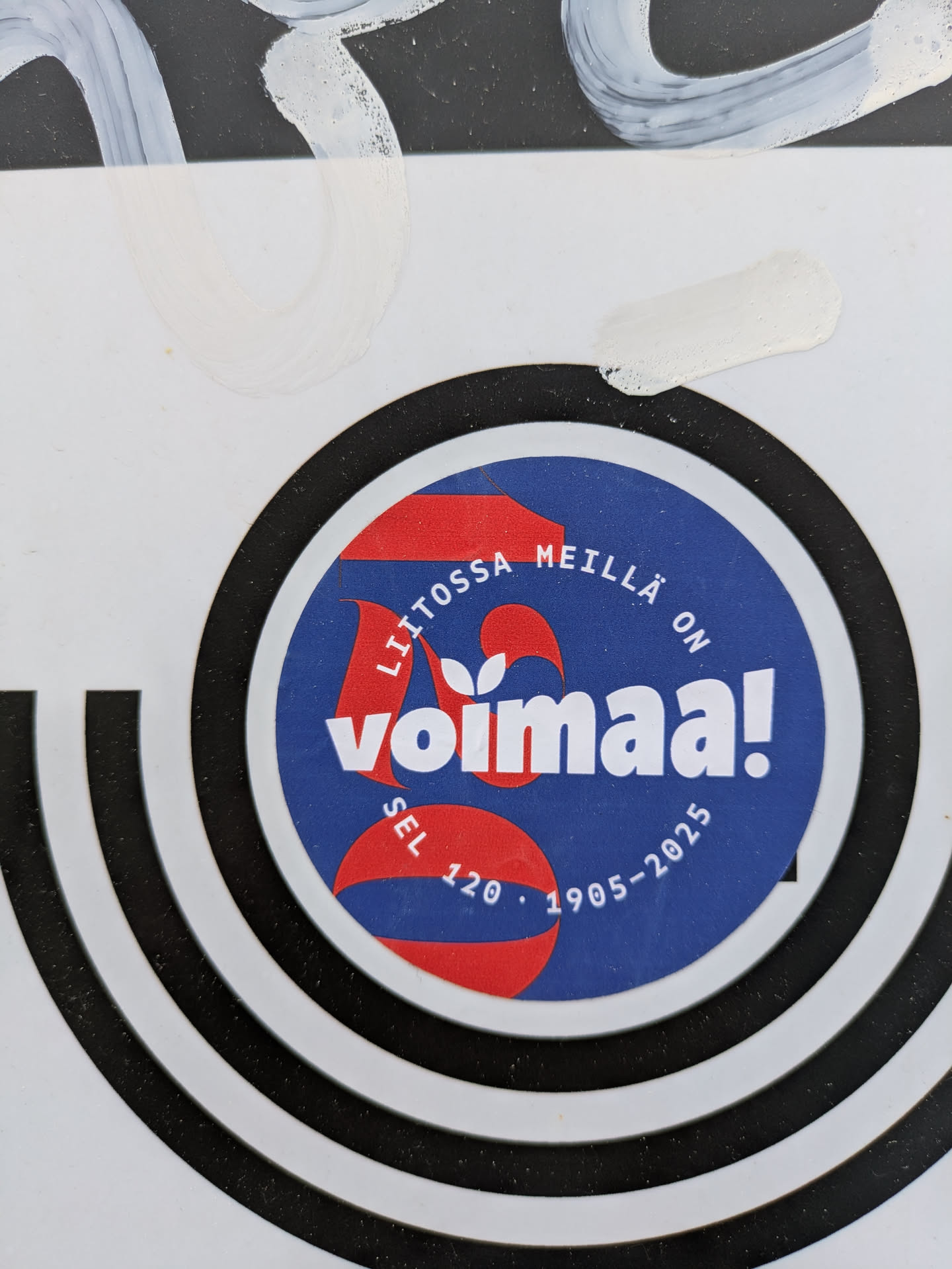 The sticker features the text "LIITOSSA MEILLÄ ON Voimaa!" in white letters on a blue background, with "SEL 120. 1905-2025" at the bottom. The word Voimaa! is stylised with a plant shoot coming out of the letter V and underneath the word is a red rounded rectangular shape.  The sticker is round and is surrounded by a black ring, which in turn is surrounded by a white background with black stripe design. The sticker is stuck on what looks like a wall and some white paint is visible on the top part of the image.