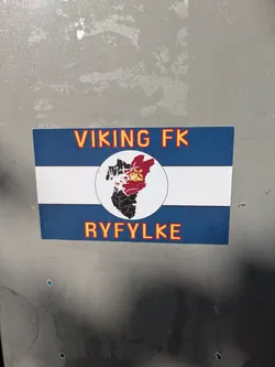 The sticker is rectangular with a design resembling a flag, featuring a wide blue band at the top, a white horizontal stripe in the middle, and another wide blue band at the bottom. In the center of the white stripe, there's a circular emblem. This emblem contains a stylized map or geographic outline, with parts colored red and black, and a yellow symbol (possibly a crown or emblem) on the red section. Above the central emblem, the text 'VIKING FK' is written in orange-yellow letters with a red outline. Below the emblem, 'RYFYLKE' is written in the same style. The overall design suggests support for the football club Viking FK and its connection to the Ryfylke region.