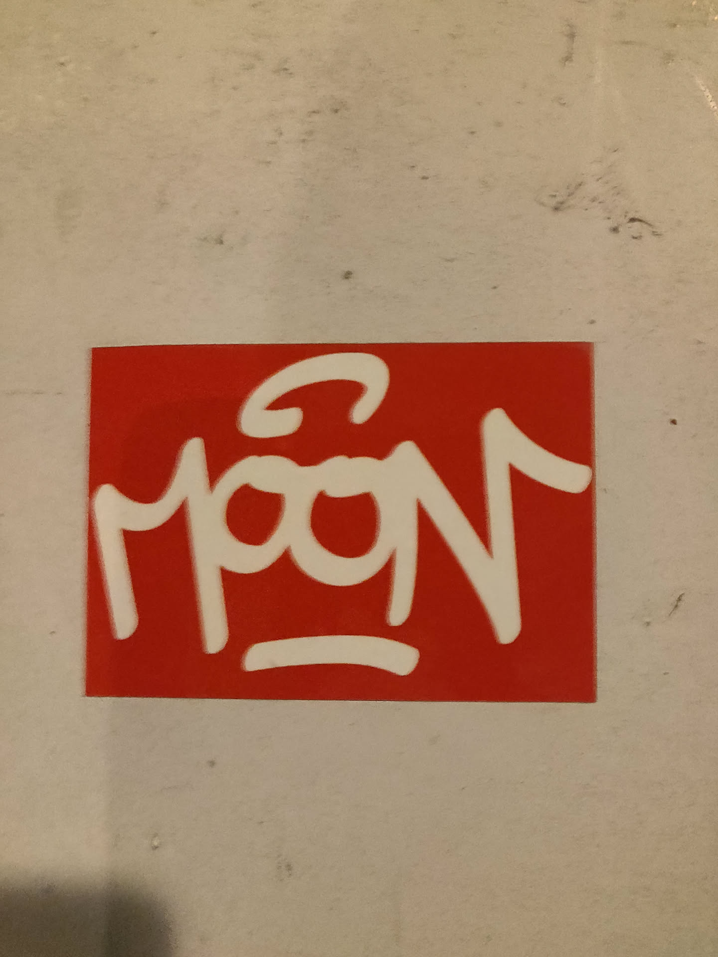 A square red sticker with the word "MOON" written in white, stylized graffiti lettering. The "M" has a unique flourish above it.  The sticker appears to be made of vinyl or similar material and is adhered to a light beige surface.