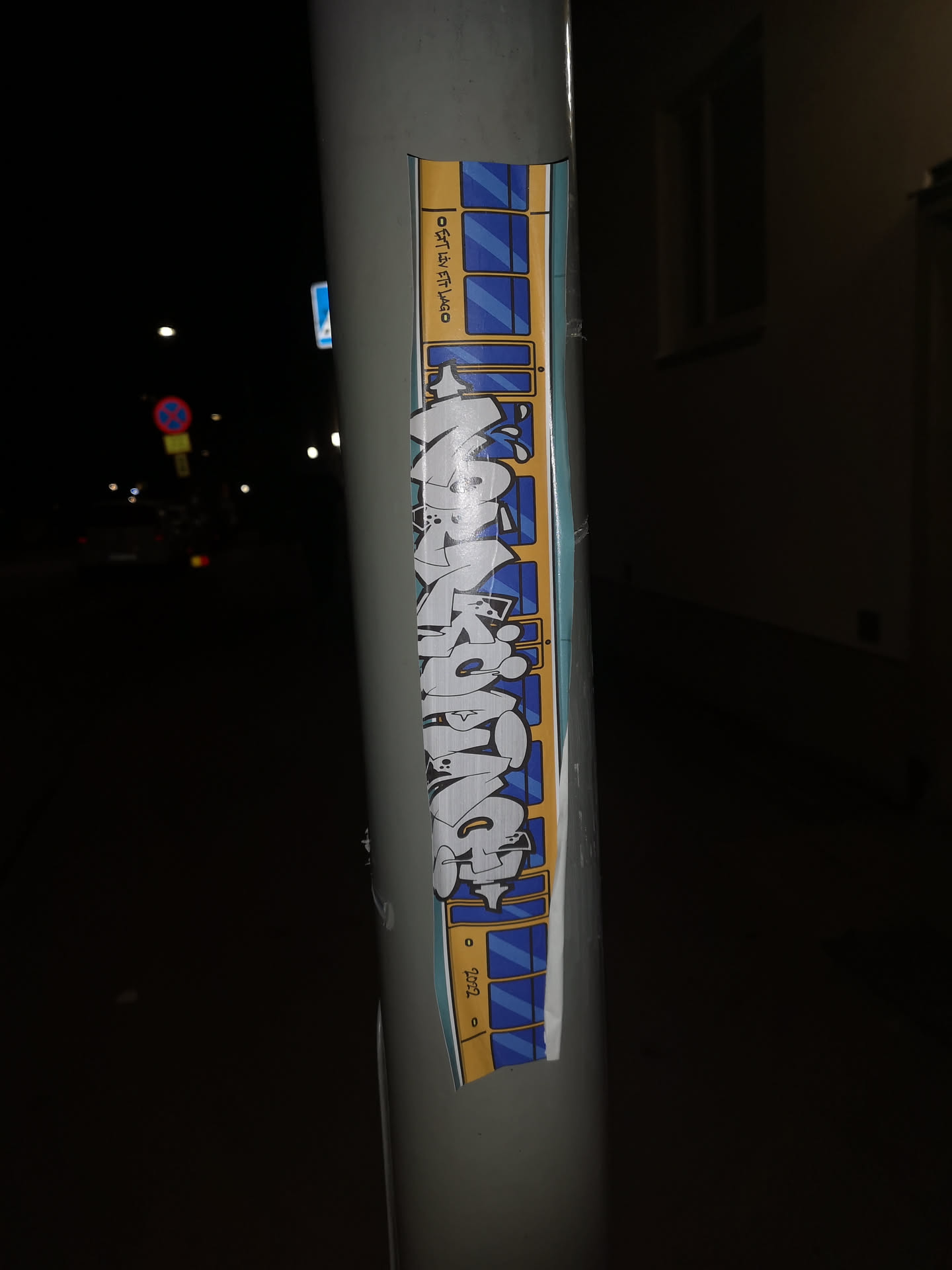 The sticker is a vertical, rectangular design depicting a stylized train car. It features a yellow top and bottom border, with a light blue central band containing multiple rectangular windows with dark blue panes, simulating the side of a train. Overlaid on this train car background is a large, intricate graffiti tag in a 'bombing' or 'wildstyle' aesthetic. The tag, which spells 'MAZKOMA', is primarily white with grey and light blue shading, outlined in black. The letters are bubbly, interconnected, and have a dynamic, three-dimensional appearance. Small details like drips or splatters are incorporated into the lettering. The sticker is applied to a grey pole, showing slight wear and a minor wrinkle at the bottom.