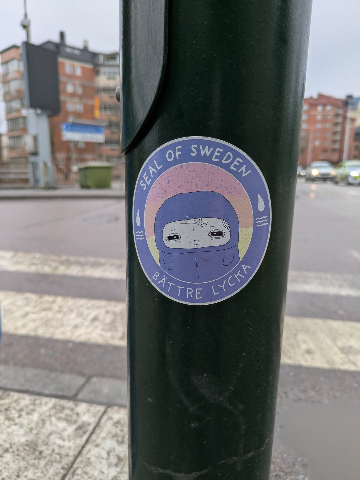 A round sticker is affixed to a green pole. The sticker features the text 'SEAL OF SWEDEN' in an arc at the top. Below this, there is an illustration of a character with a pale face and dark eyes, wearing what appears to be a blue hooded garment. The character has a somewhat sad or melancholic expression. The hood is colored in a shade of blue, with a hint of purple around the face. The backdrop consists of a pale orange to pink gradient. There are stylized droplets and wave symbols to the sides of the hooded figure. The text 'BÄTTRE LYCKA' is located at the bottom of the sticker.