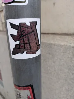 Close-up image of a square sticker featuring an abstract geometric design in shades of brown and beige. The style is reminiscent of minimalist or cubist artwork. The sticker is affixed to a gray metal pole. 