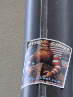 Sticker featuring a cartoon illustration of a bearded man in red and white striped sleeves holding boxes of fireworks.  The background includes an American flag and what appears to be a fireworks warehouse. The website www.PyroGiant.com is prominently displayed.