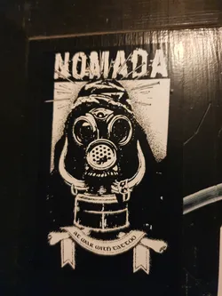 Black and white sticker featuring a figure wearing a gas mask with a third eye, surrounded by pins. The text 'Nomadada' is at the top, and a banner at the bottom says 'at war with tattoo'.