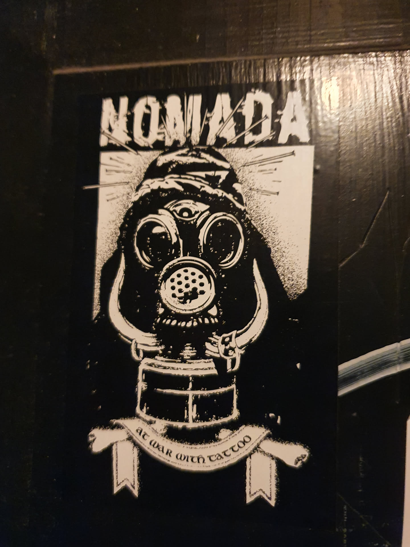 Black and white sticker featuring a figure wearing a gas mask with a third eye, surrounded by pins. The text 'Nomadada' is at the top, and a banner at the bottom says 'at war with tattoo'.