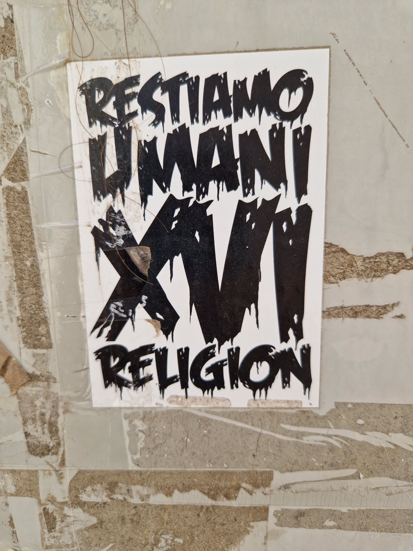 A black and white sticker with dripping text. The text says "Restiamo Umani XVI Religion". The sticker is attached to a wall.
