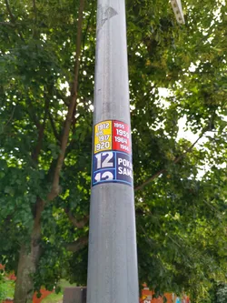 A sticker featuring different years (1912, 1915, 1917, 1920, 1955, 1959, 1964, 1966) and the numbers 12 and 13 with the partially visible words 'POKA' and 'SAML'.  It's affixed to a metal lamppost against a backdrop of lush green trees.