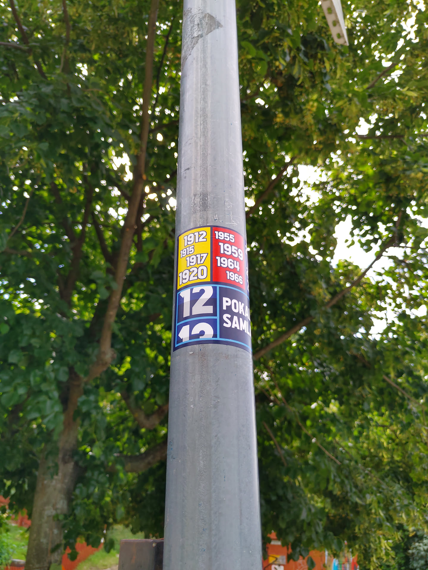 A sticker featuring different years (1912, 1915, 1917, 1920, 1955, 1959, 1964, 1966) and the numbers 12 and 13 with the partially visible words 'POKA' and 'SAML'.  It's affixed to a metal lamppost against a backdrop of lush green trees.