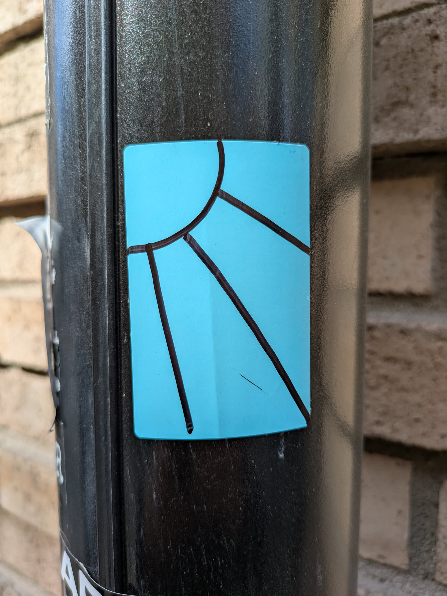 The sticker features a simple design of a sun with rays, drawn with black lines on a turquoise background. It is affixed to a black pole. The design suggests a sunny and vibrant theme.