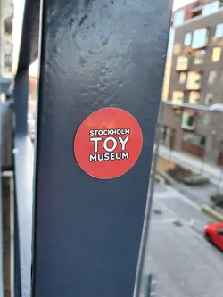 A red circular sticker with the text "STOCKHOLM TOY MUSEUM" in white. It's affixed to a dark gray metal pole outdoors.