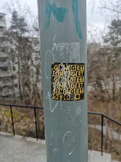A rectangular sticker with a yellow and black checkerboard or grid pattern, affixed to a grey metal pole. The word 'GNAGET' is prominently displayed multiple times across the grid in black capital letters on yellow squares, and possibly yellow letters on black squares, though much of the sticker is obscured by white paint or marker streaks. Below the main text, there is a row of five small, distinct icons or emblems. From left to right, these include a crest-like symbol, a stylized face or crown, a circular emblem with text, a sunburst design, and another shield-like badge. The sticker shows signs of wear and tear, with parts of its surface scratched or faded.
