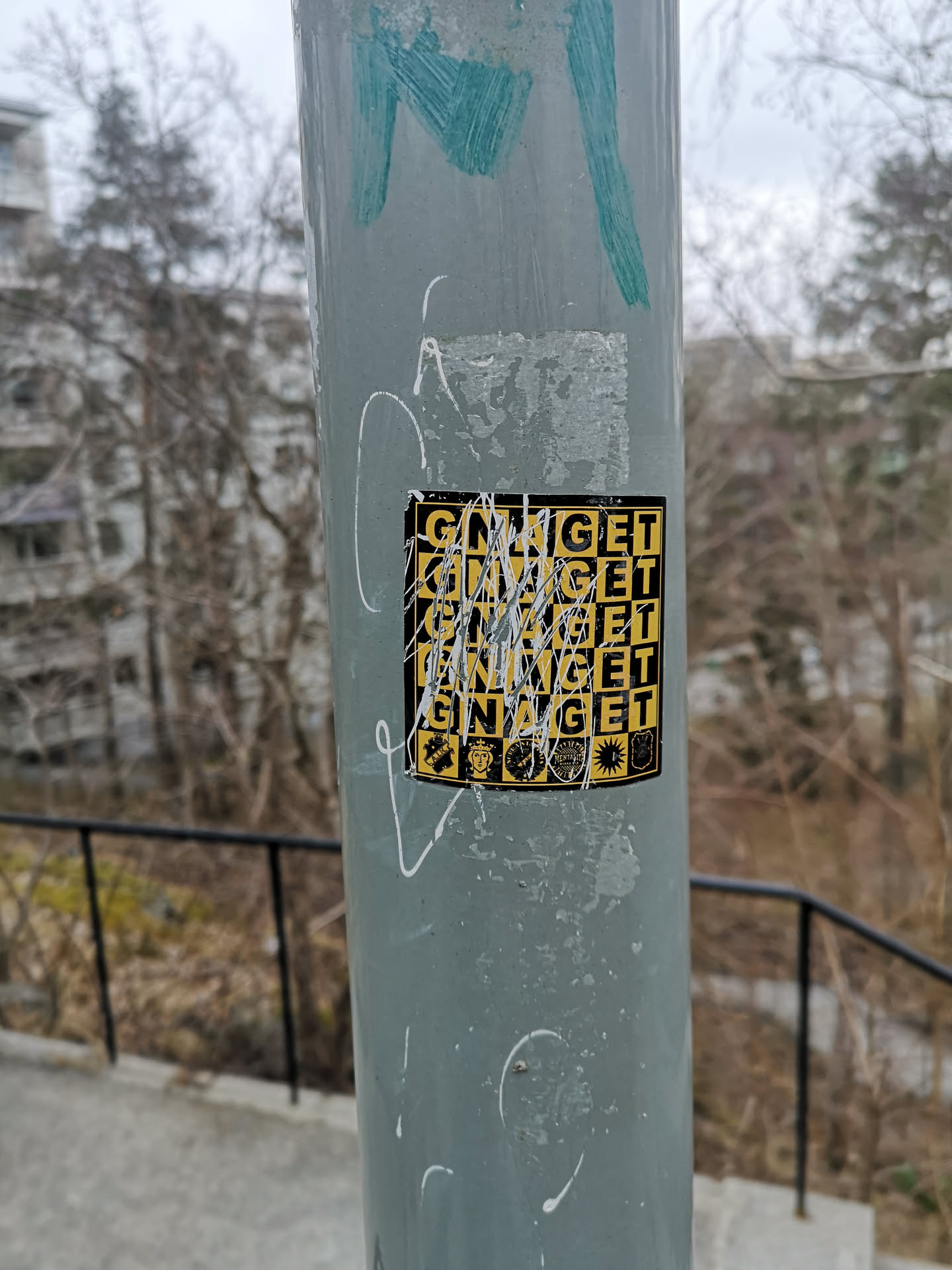 A rectangular sticker with a yellow and black checkerboard or grid pattern, affixed to a grey metal pole. The word 'GNAGET' is prominently displayed multiple times across the grid in black capital letters on yellow squares, and possibly yellow letters on black squares, though much of the sticker is obscured by white paint or marker streaks. Below the main text, there is a row of five small, distinct icons or emblems. From left to right, these include a crest-like symbol, a stylized face or crown, a circular emblem with text, a sunburst design, and another shield-like badge. The sticker shows signs of wear and tear, with parts of its surface scratched or faded.