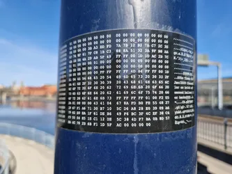 A sticker with a long sequence of hexadecimal code is attached to a blue pole.  Some text is visible on the right side of the sticker, possibly containing notes or metadata related to the code. The surrounding environment appears to be outdoors, near water.