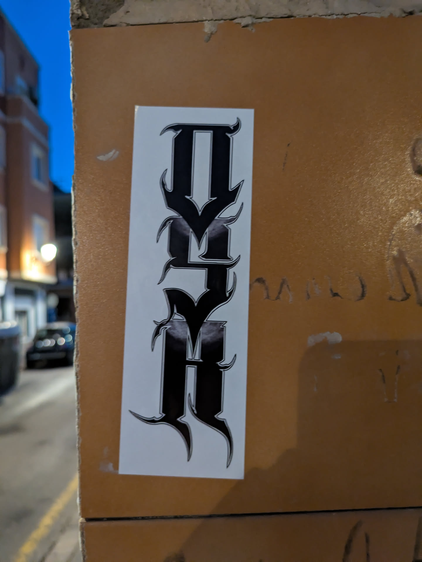 The image features a white sticker affixed to a textured, orange-brown surface. The sticker displays a stylized word or abbreviation stacked vertically with black lettering. The letters are ornate, with pointed edges and a gothic or metal-inspired design. The text appears to read 'THVR' or 'THYR'.