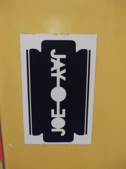 A black and white sticker of a razor blade with the text "JOE JAX" in the center. The sticker is on a yellow surface.