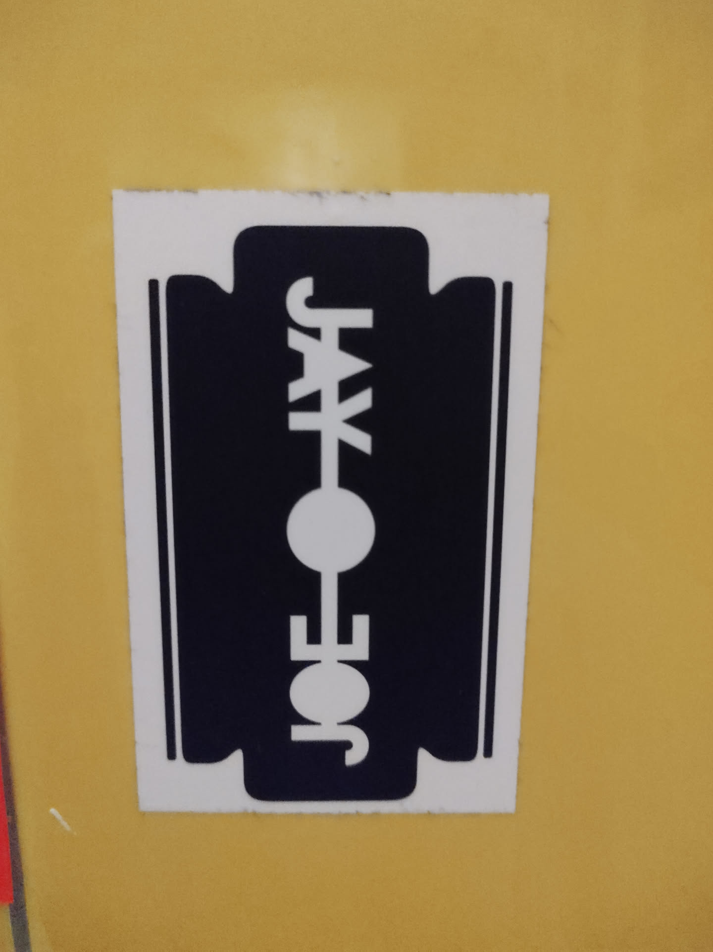 A black and white sticker of a razor blade with the text "JOE JAX" in the center. The sticker is on a yellow surface.