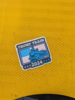 The sticker features a stylized blue steam train with 'TRUMP TRAIN' emblazoned above it in white text on a dark blue background with a white border. Behind the train is a representation of the American flag, with its stars and stripes. Below the train, '2024' is displayed, surrounded by several small blue stars.