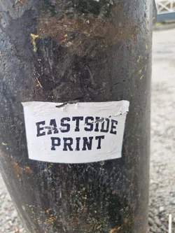 A simple sticker with the text "Eastside Print" in a dark-colored, sans-serif font on a white background affixed to a dark-colored pole.